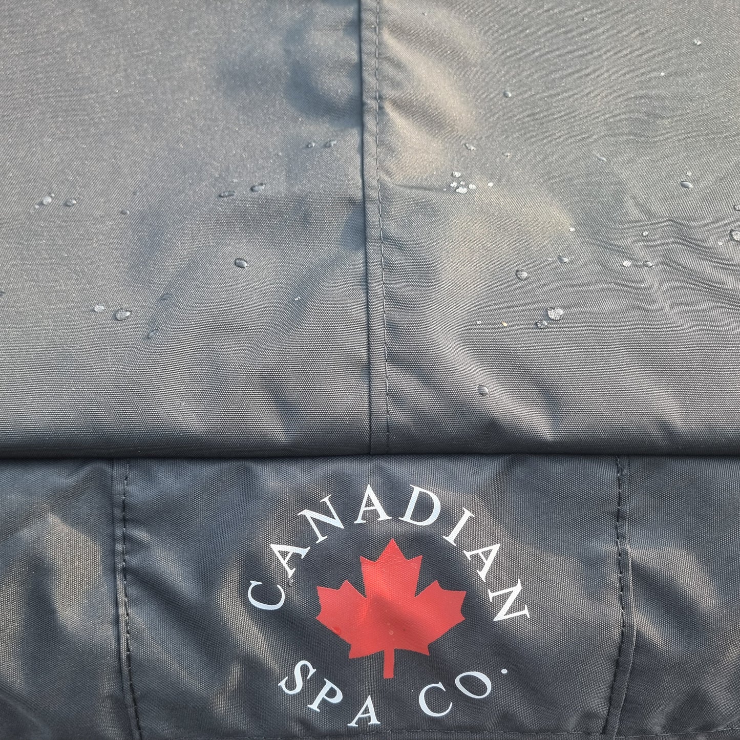 Close-up of a black oxford material that is waterproof fabric with water droplets on the surface and a stitched Canadian Spa Co. logo featuring a red maple leaf in the lower section.