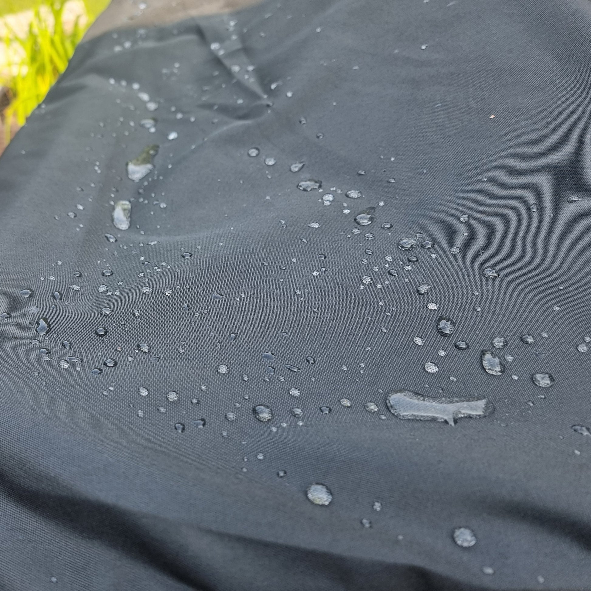 A close-up of a black oxford  waterproof fabric, likely a hot tub cover, with water droplets beaded on its surface. The background shows a blurred outdoor patio setting with a wooden bench and greenery.