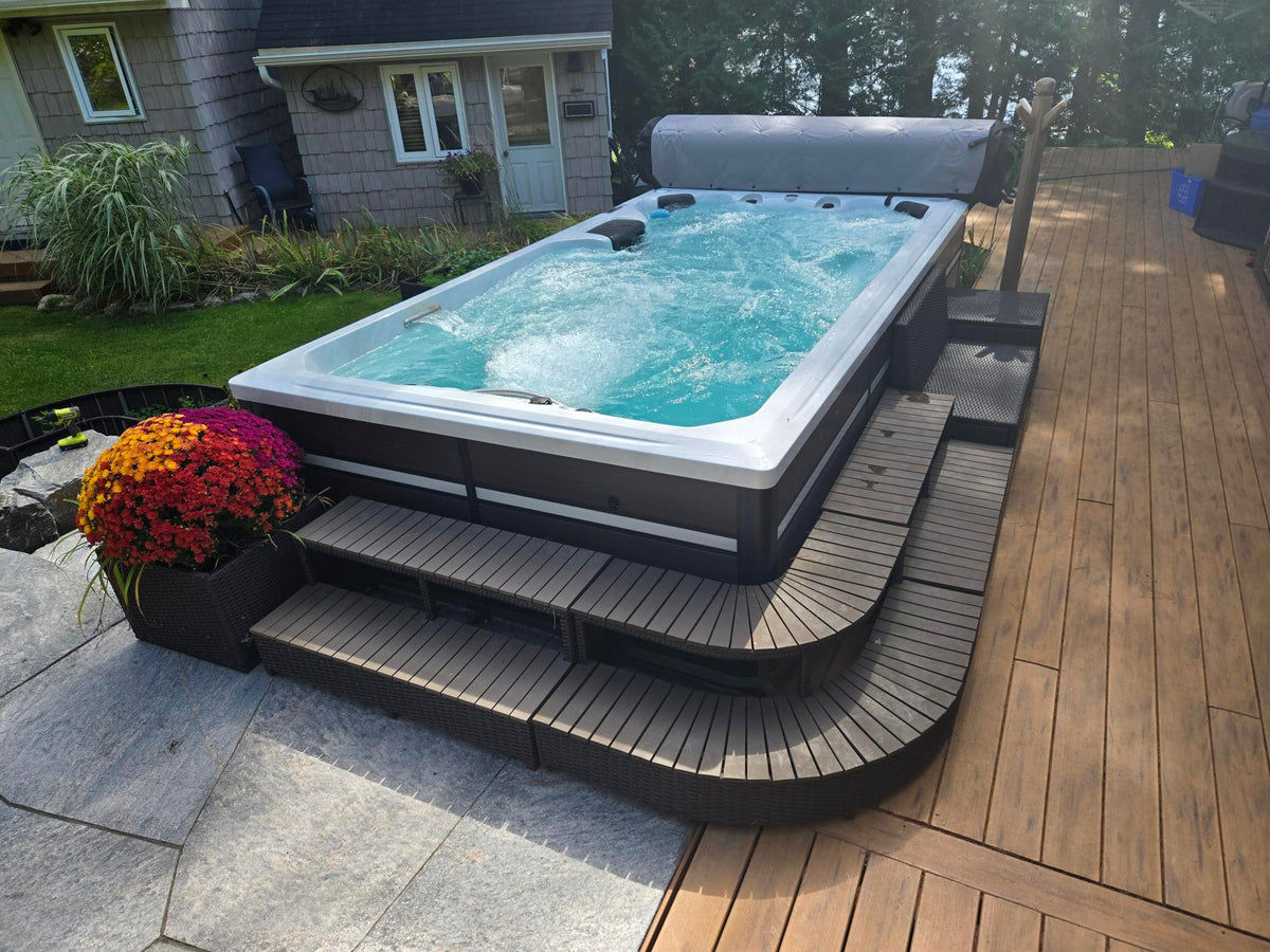 An outdoor hot tub with bubbling turquoise water, set on a curved dark wood platform next to a wooden deck and house yard.